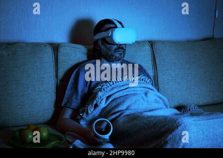 L'uomo che utilizza il visore per realtà virtuale guarda la TV o il film al buio, la persona seduta sul divano guarda gli occhiali VR. Concetto di gioco di videogiochi, futuristico Foto Stock