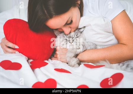 Donna sdraiata sul letto con cuori rossi e baciando il suo bel gatto. Amore per gli animali domestici. Concetto di San Valentino Foto Stock
