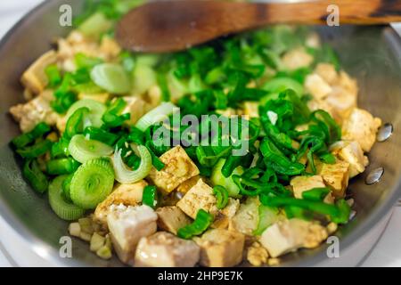 Tofu cubetti frittura in teglia inox con cipolla verde tritata negi verdure e spatola di legno macro closeup che mostra la consistenza in cucina sto Foto Stock