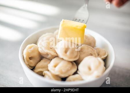 Primo piano macro di gnocchi tradizionali russi pelmeni con ripieno di carne in ciotola bianca con forchetta a mano che mette il burro su cibo caldo in tradizione Foto Stock