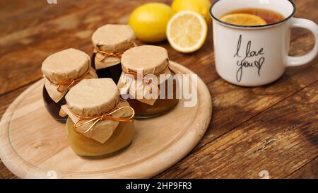 Colazione dolce. Tazza di tè nero con limone. Limoni e quattro vasetti con marmellata fatta in casa, marmellata o cagliata su un tavolo di legno. Confit di frutta Foto Stock