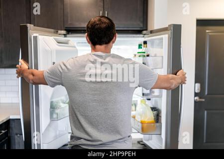 Retro di uomo affamato aprire frigorifero porte elettrodomestici frigorifero alla ricerca di cibo all'interno con condimenti e succo di frutta in cucina moderna Foto Stock