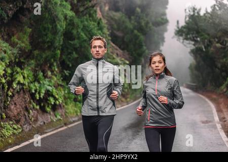 Coppia in corsa sul sentiero parco bagnato jogging in pioggia indossando giacche sportive in climi freddi. Donna asiatica, uomo caucasico atleti allenamento cardio Foto Stock