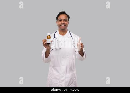 L'uomo indiano Dottore che tiene sciroppo di tosse che mostra Thumb in su isolato Foto Stock
