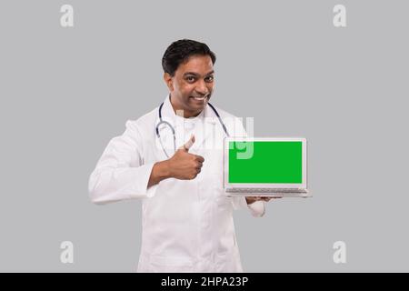 Il medico mostra il pollice su con schermo verde del portatile isolato. Medico indiano maschio con portatile in mani. Foto Stock