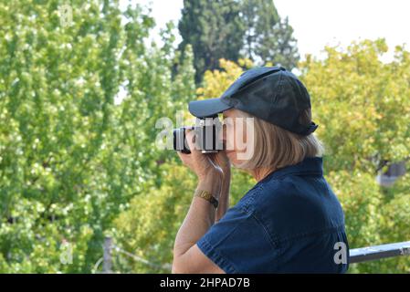 Una donna anziana fotografo che utilizza una fotocamera reflex 35mm a livello degli occhi. Abito blu e cappuccio su un morbido sfondo di alberi Foto Stock