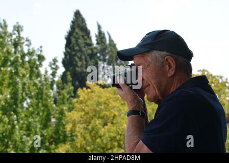 Fotografo maschile anziano che utilizza una fotocamera SLR da 35mm. Maglietta blu con cappuccio blu su un morbido sfondo di alberi verdi. Foto Stock