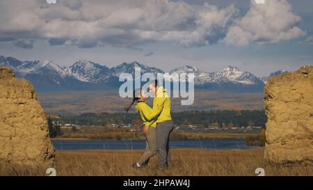 Uomo e donna in abbigliamento sportivo giallo verde. Una coppia incantevole di viaggiatori si abbraccia e bacia vicino alla pietra vecchia godendo il paesaggio delle Highland. Due viaggiatori camminano sullo sfondo di montagne innevate. Foto Stock
