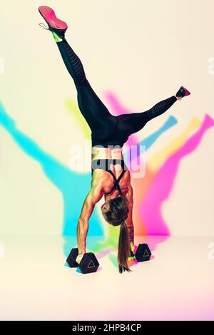 La donna forte fa il handstand di esercitazione gli spintups sui dumbbells. Foto di donna atletica in nero sportswear su sfondo bianco con effetto di colori rgb s. Foto Stock