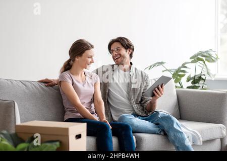 Felice felice giovane caucasico marito in occhiali e moglie in casual relax sul divano con tablet, progettazione di nuovi interni Foto Stock
