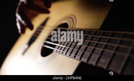 Il musicista ama suonare la chitarra acustica seduta nella stanza buia. Mano dell'esecutore che tocca le corde dello strumento musicale su sfondo nero vista ravvicinata Foto Stock