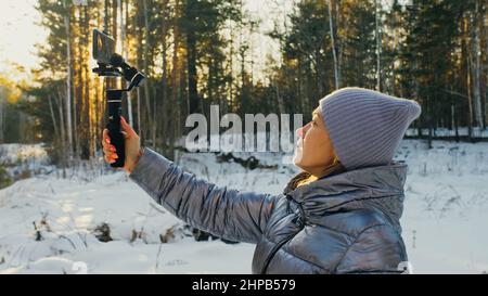 Il videografo professionale donna che tiene smartphone su 3-asse dispositivo di stabilizzazione gimbal in inverno. Pro Equipment aiuta a realizzare video di alta qualità al telefono. Operatore cinematografico. Movimento lento Foto Stock