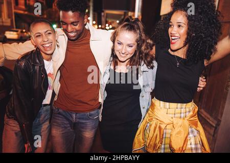 Uscire il venerdì sera. Gruppo di giovani vivaci che ridono felicemente mentre camminano insieme in città di notte. Amici multiculturali che hanno Foto Stock