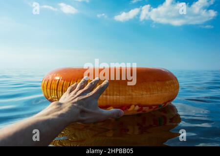 La mano di una persona si allunga per aiutare un cerchio gonfiabile multicolore per nuotare sull'acqua di mare in una calda giornata di sole, un galleggiante di ciambella per nuotare Foto Stock