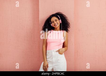 Bella donna sorridente che indossa abiti casual luminosi appoggiando parete rosa ascoltare musica Foto Stock