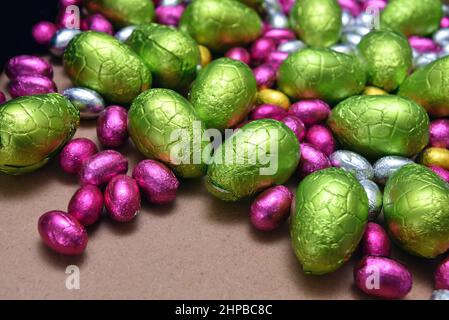 Un piccolo foglio rosa, giallo, verde e argento avvolse uova di pasqua al cioccolato con uova verdi di calce più grandi, su un fondo di legno chiaro. Foto Stock