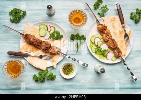 Due porzioni di appetitoso kebab shish fatto in casa, cetrioli e ravanelli tagliati a fette, bicchieri di vino vista dall'alto su sfondo blu di legno. Foto Stock