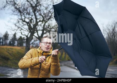Uomo che tiene rotto ombrello in forte vento durante la notte di pioggia cupa. Temi meteo e meteorologia. Foto Stock