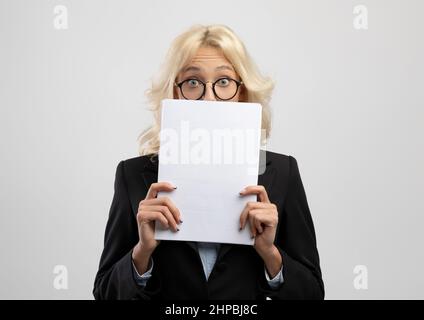 Scioccato imprenditore femmina che sbircia da dietro documenti, posando su sfondo grigio chiaro studio. Sorpreso segretario in occhiali guardando attraverso Foto Stock