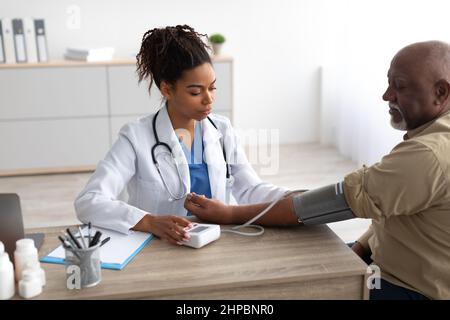 Ipertensione in età avanzata. Giovane lavoratore medico femminile che misura la pressione arteriosa dell'uomo nero anziano usando il bracciale, paziente che ha problemi con Foto Stock