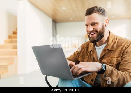 Maschio entusiasta che utilizza gli auricolari indossati da notebook che lavorano online a casa Foto Stock