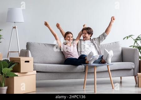 Felice giovane caucasico marito e moglie in casual alzare le mani in su festeggiando muoversi sul divano Foto Stock