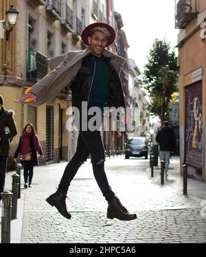 Modello giovane di moda con giacca grigia e cappello rosso che salta per strada. Foto Stock