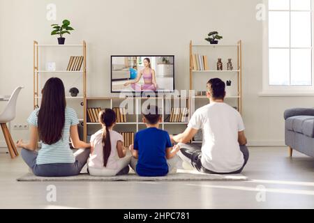 La giovane famiglia con bambini pratica lo yoga e medita insieme a casa davanti allo schermo della TV. Foto Stock