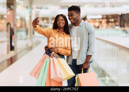 Buoni sposi neri che acquistano nel centro commerciale moderno della città e prendono Selfie sullo smartphone, coppia afro-americana che ha divertimento, godendo fare gli acquisti Toge Foto Stock