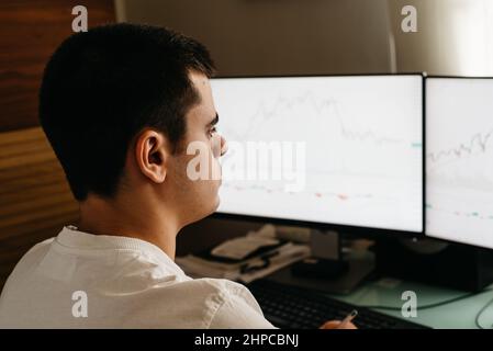 Giornata di lavoro a casa. Vista laterale del commerciante giovane riuscito nell'abbigliamento casuale che lavora con le tabelle e le relazioni di mercato sugli schermi del calcolatore nella sua sede. Foto Stock