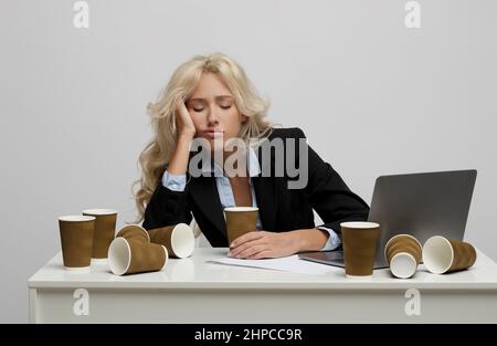 Lavoratore di ufficio femminile esausto circondato da tazze di caffè vuote che dormono sul posto di lavoro su sfondo grigio chiaro studio Foto Stock