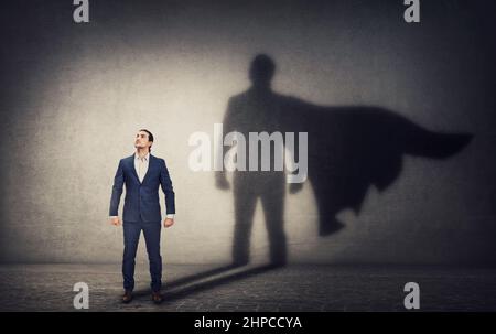 Un uomo d'affari determinato è fiducioso in una posizione di eroe e getta un'ombra coraggiosa del supereroe sul muro dietro. Leadership aziendale e motivazione c Foto Stock