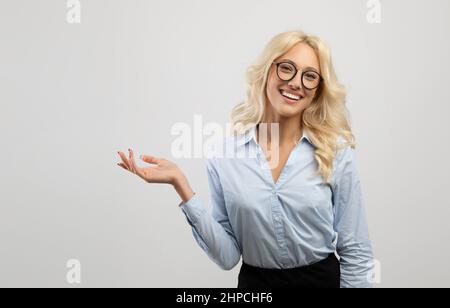 Offerta business. Felice donna d'affari bionda che dimostra qualcosa al suo palmo vuoto, tenendo un oggetto invisibile e sorridendo alla macchina fotografica sul backgro grigio Foto Stock