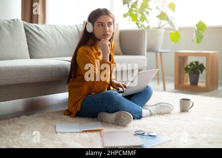 Videochiamata online. Giovane donna premurosa in cuffie sedute sul pavimento, utilizzando un computer portatile, con conferenza virtuale al chiuso Foto Stock