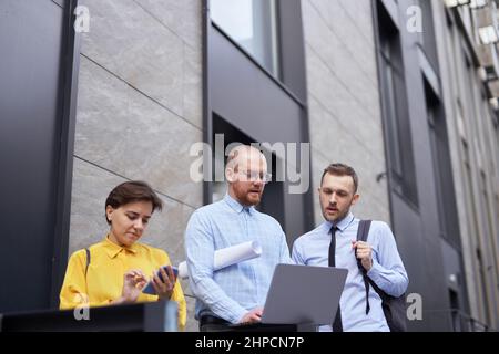 Uomini e donne in abiti casual in piedi all'esterno, utilizzando un computer portatile, parlando e avendo brainstorming o discutere di progetto. Team di persone d'affari che guardano e discutono la presentazione del progetto all'aperto Foto Stock