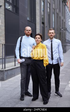 Bella donna caucasica fiduciosa in blusa gialla e due uomini che guardano la macchina fotografica sorridendo. Ritratto medio di successo del team professionale di affari che si pone all'aperto nel centro città. Foto di alta qualità Foto Stock