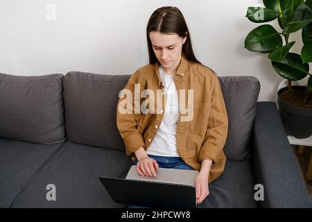 Premuroso interessato giovane donna seduta su un comodo divano di lavoro sul computer portatile, pensare risolvere il problema a casa, grave caucasica femmina 20s ricerca fo Foto Stock