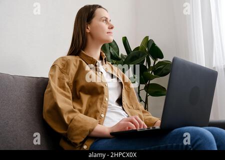 Premuroso giovane donna 20s seduta sul divano con laptop pensando di ispirazione sognando guardando lontano profondo perso nei pensieri, sognante ragazza brunette relax Foto Stock