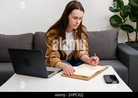 Giovane ragazza studentesca adulta seduta sul comodo divano nel soggiorno con chiamata virtuale online, webinar di chat educativa a casa, scrivendo appunti. Foto Stock