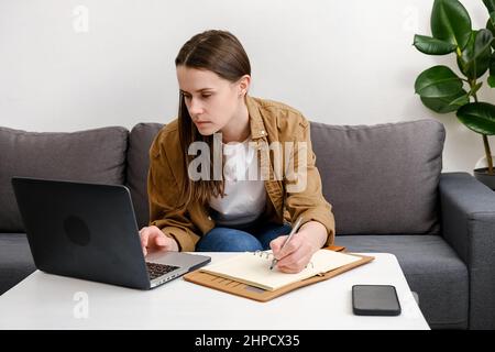 Una giovane donna focalizzata si siede sul divano usando il laptop a casa, guardando lo schermo, chiacchierando, leggendo o scrivendo e-mail Foto Stock