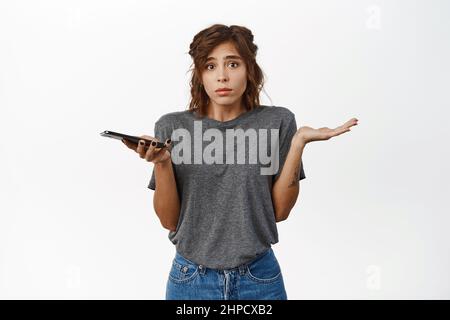 Giovane donna confuso con smartphone, spalle e tenere telefono cellulare, non so, essere indecisa, in piedi su sfondo bianco Foto Stock