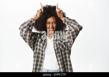 Felice e sassosa giovane donna africana mostra corna di toro, dita sul gesto della testa e sorridente, posando allegro e spensierato, in piedi su sfondo bianco Foto Stock