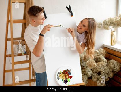 Giovane coppia carino stand dietro cavalletto con tela bianca e tavolozza toccare con pennelli vicino scala di legno e fiori. Foto Stock