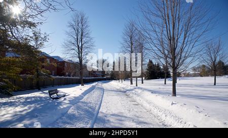 Vista di un quartiere residenziale con un sentiero nel parco con neve profonda e una panchina parco. Foto Stock