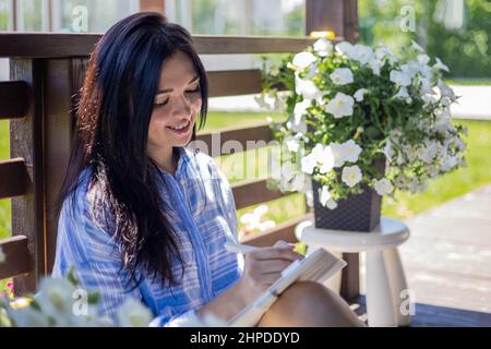 Giovane donna asiatica pensiva che scrive diario di carta seduta sulla terrazza estiva all'aperto in campagna Foto Stock
