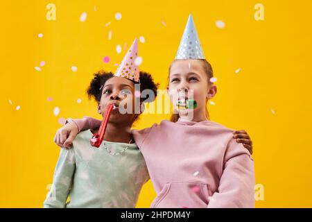 Ritratto di due ragazze che indossano cappelli da festa mentre festeggia compleanno su sfondo pop giallo in studio Foto Stock