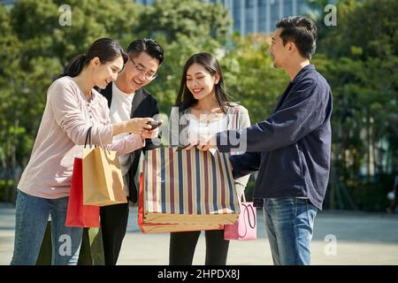 gruppo di quattro giovani asiatici che guardano il cellulare che parlano durante il viaggio di shopping felice e sorridente Foto Stock