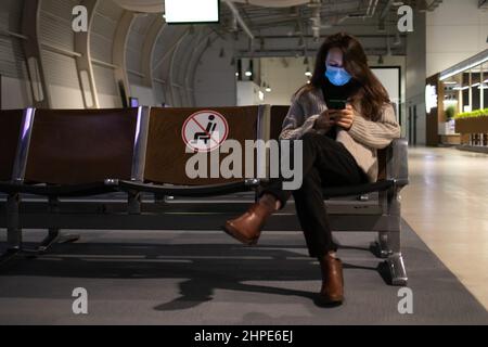 Una giovane donna in maschera medica è seduta in aeroporto e controllare il programma nel suo telefono Foto Stock