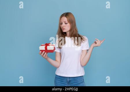 Regalo romantico. Felice giovane donna apre valentine giorno dono su sfondo blu studio, spazio copia. Bella donna che celebra una vacanza romantica, con una scatola con un nastro rosso in una maglietta bianca Foto Stock