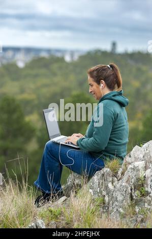 Giovane ispanica femmina in un abito sportivo seduto su rocce di montagna che lavora su un computer portatile Foto Stock